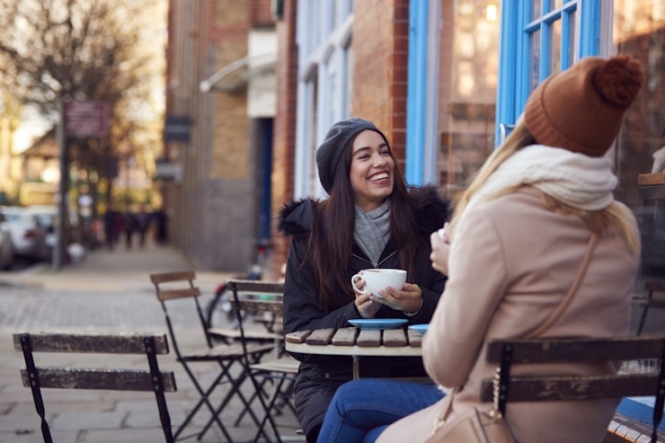 Two women wearing wooly hats sit outside with coffee cups smiling and laughing.