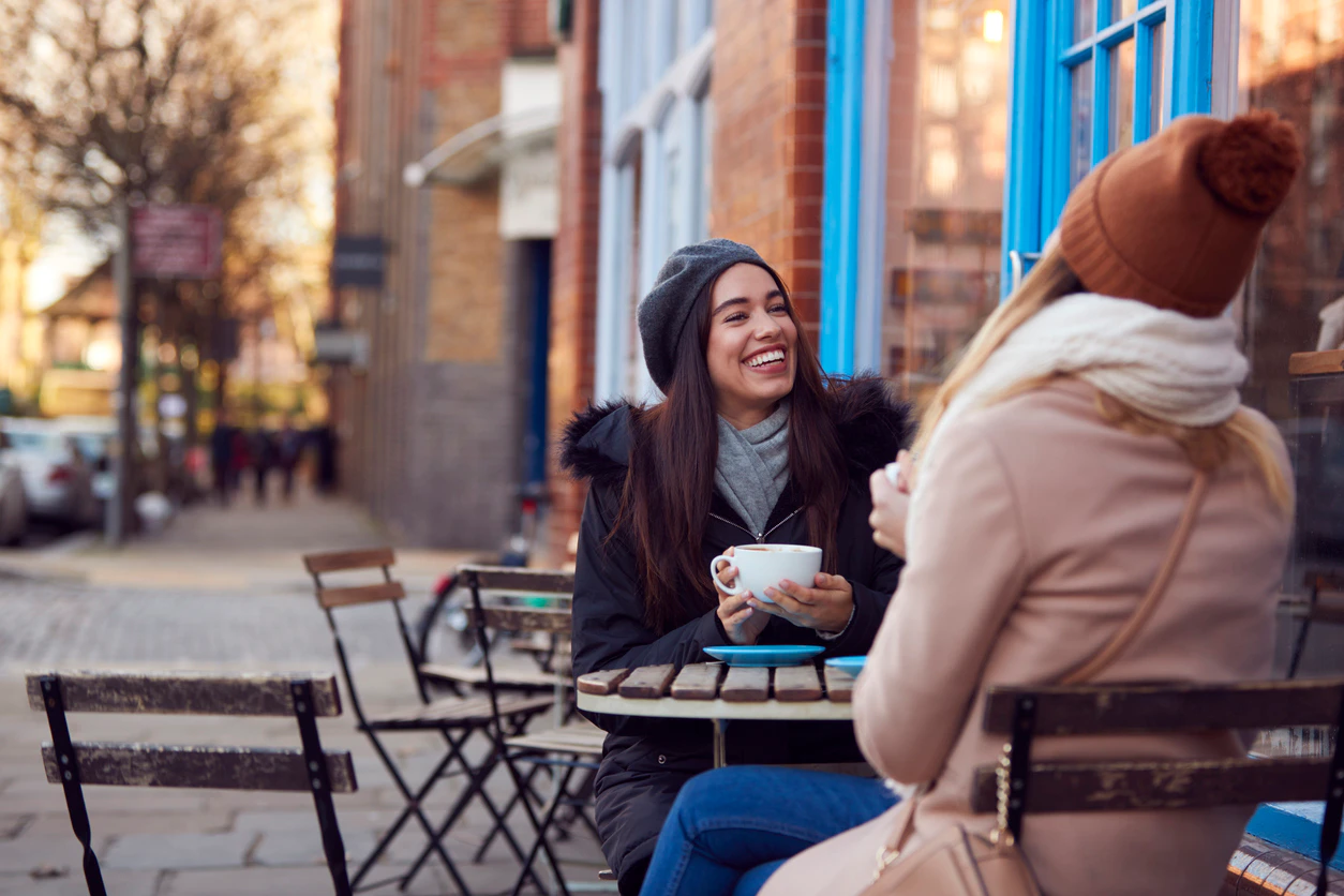 Two women wearing wooly hats sit outside with coffee cups smiling and laughing.