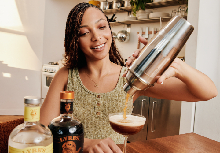 A woman with a smile on her face pours a brown non-alcoholic cocktail from a metal shaker into a martini glass.