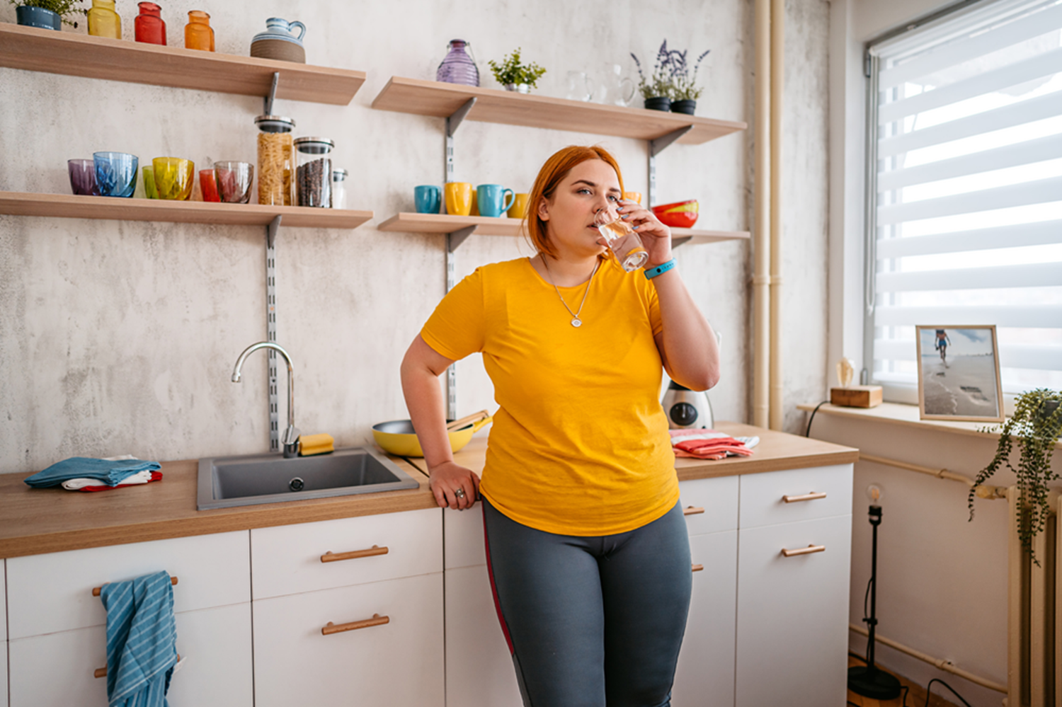 A woman wearing a yellow T-shirt drinks water in a kitchen.
