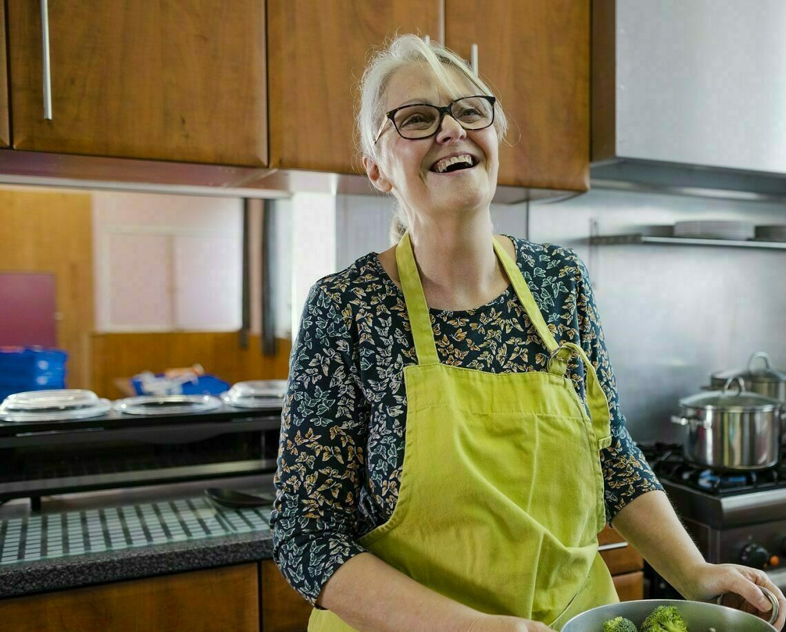 Woman wearing an apron laughs and smiles as she prepares a pan of green vegetables in a kitchen with others.