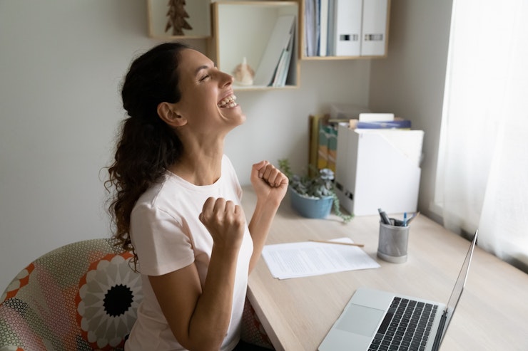 Woman with long brown curly hair clenches her fists in celebration while sitting at a desk with a laptop on it.
