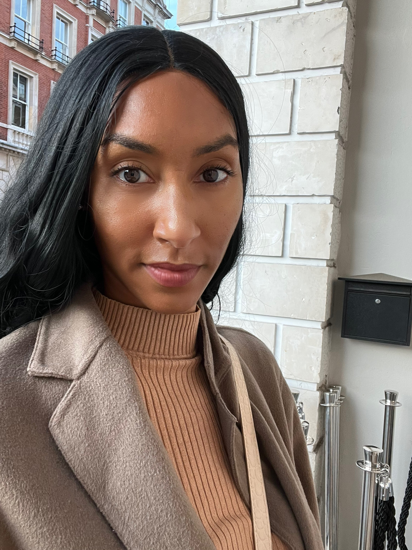 A woman with long hair wearing a collared coat sits inside in front of a wall.