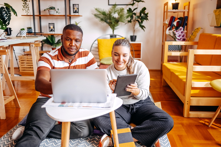 Two young people at home while sitting on floor in living room in front of laptop and tablet