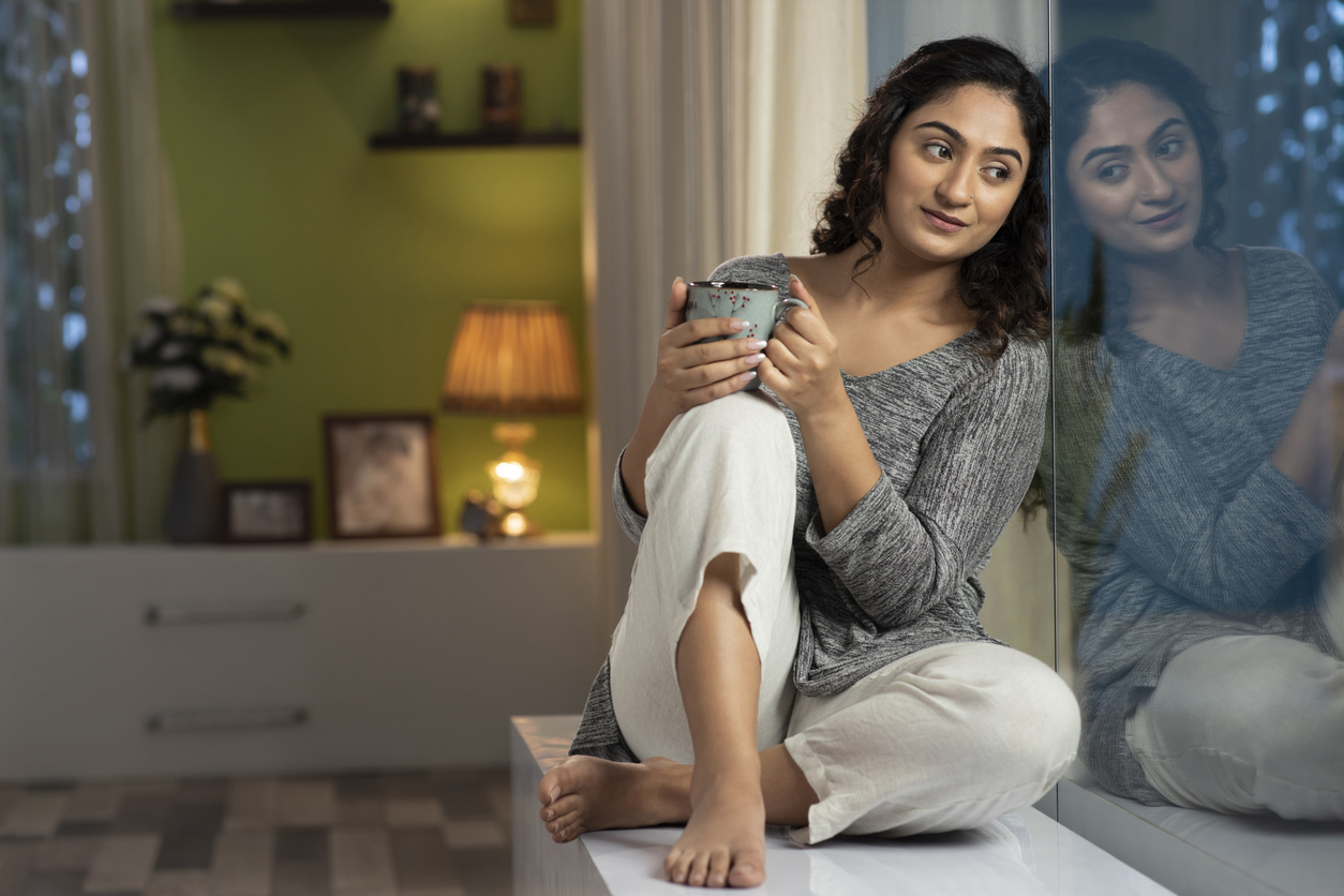 Image of young woman sitting on window sill at home drinking coffee and looking reflective