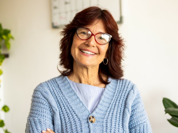 A mature woman with dark hair wearing glasses and a blue cardigan smiles broadly at the camera.