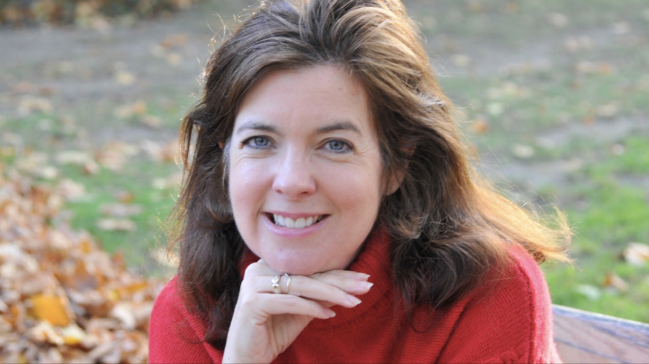 A woman with brown, shoulder-length hair rests her head on her hand and smiles from an autumnal outdoor setting.