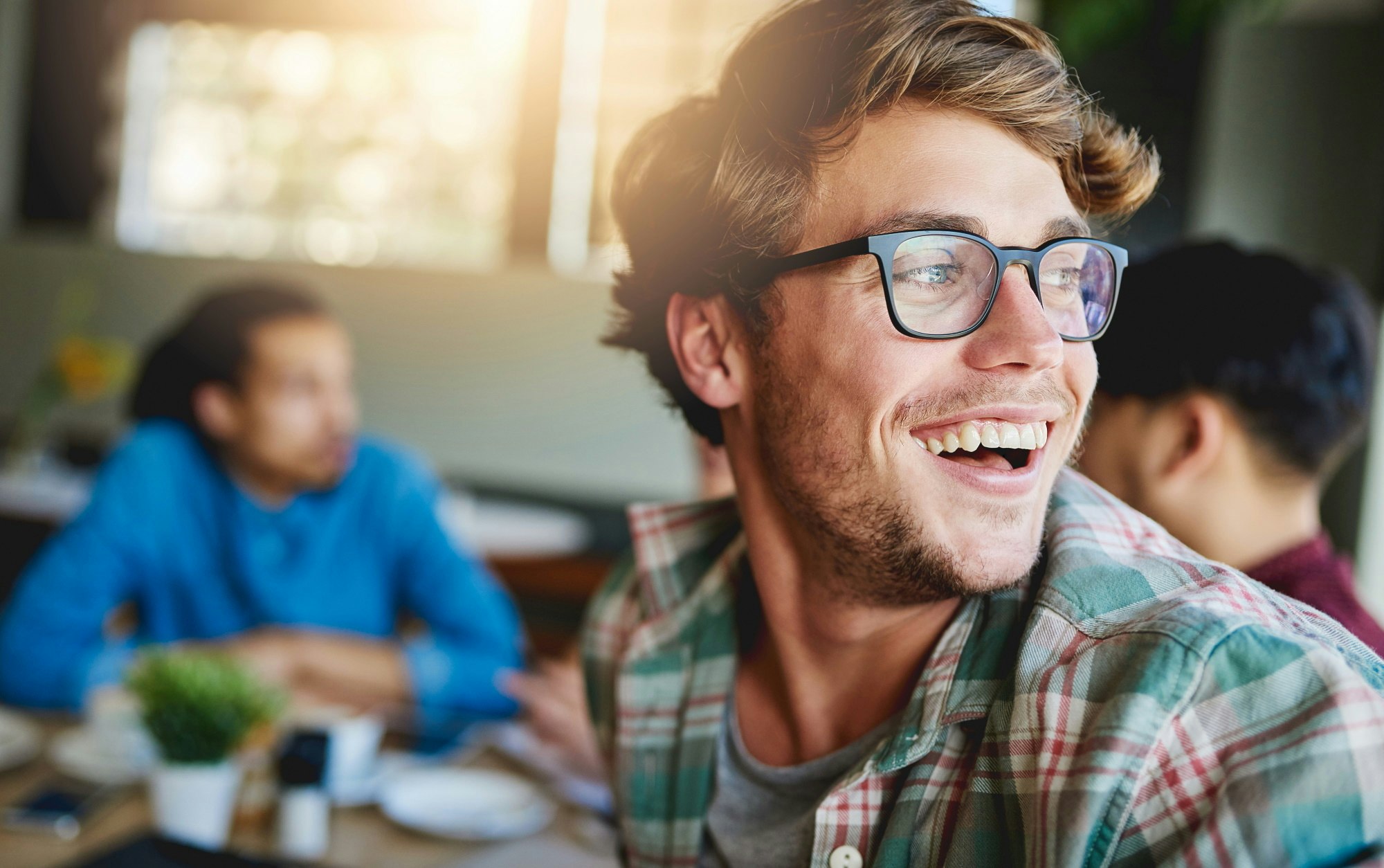 A man wearing a checked shirt smiles at a coffee shop with other people.