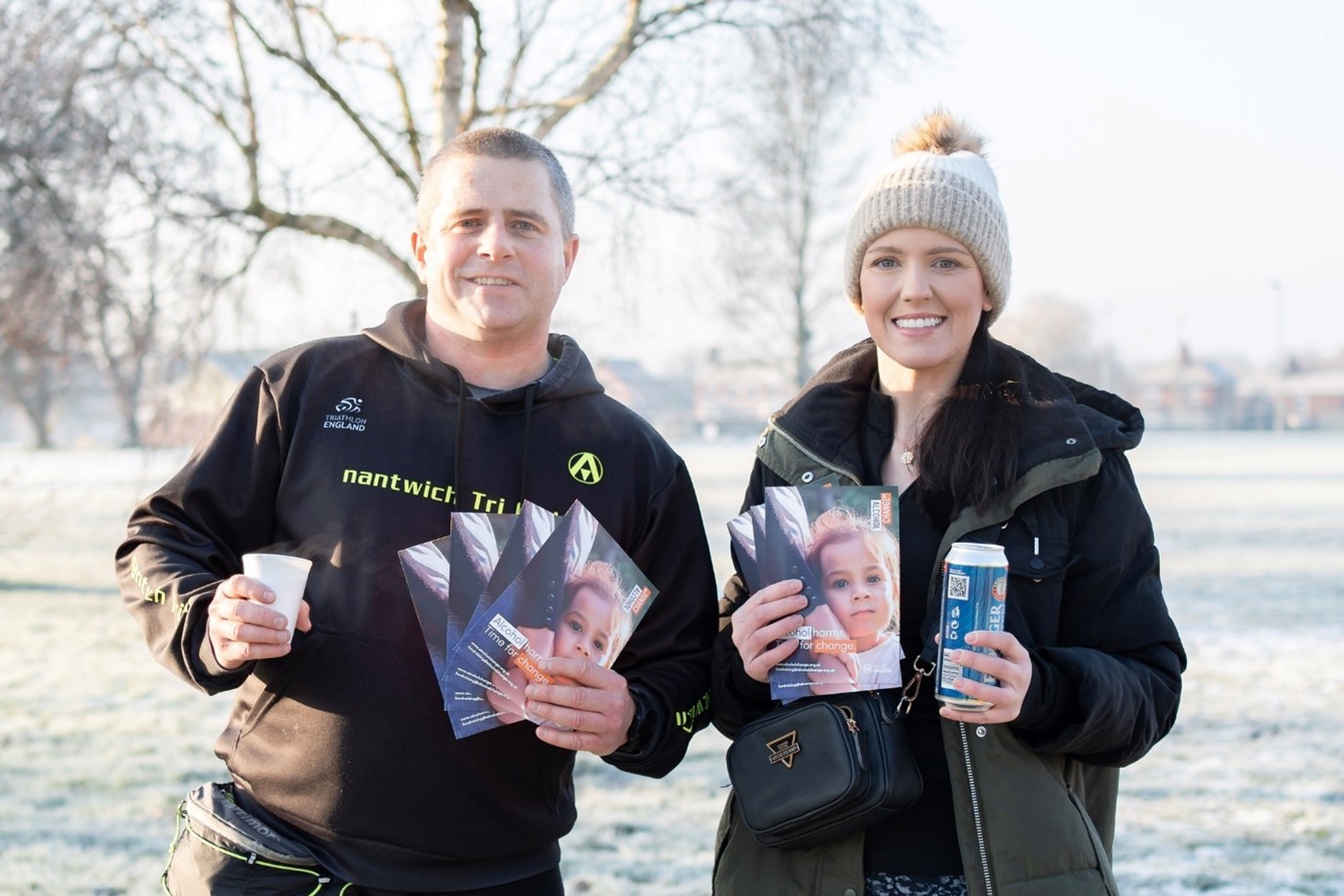 A man and a woman dressed in black winter coats hold leaflets in a wintry outdoor space.