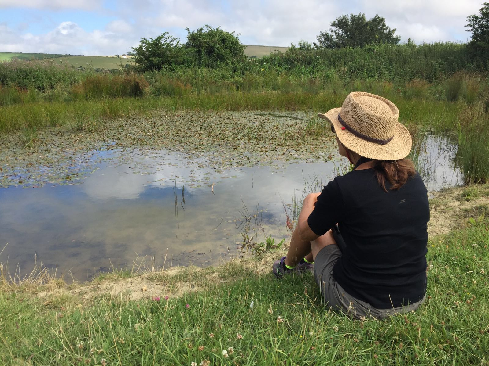 A woman wearing a straw summer hat sits on the grass of a meadow looking out at a pond.