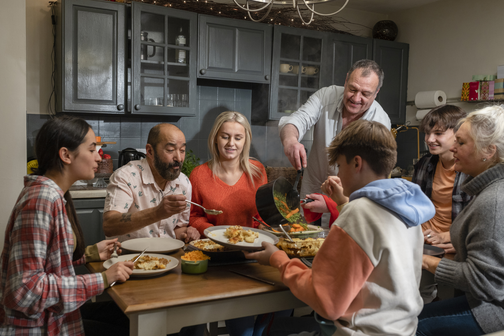 A family getting ready to have a meal together round a dining table in their kitchen with the man pouring freshly cooked carrots and peas out of a cooking pan onto a plate on the table while his family serve up the rest of the food.