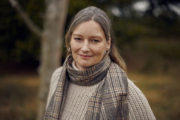 A middle-aged woman swearing a scarf and standing near woodland looks at the camera.