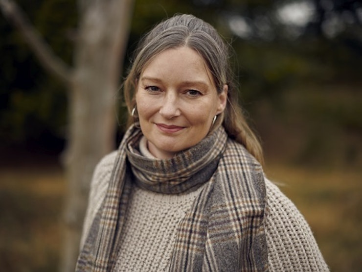 A middle-aged woman swearing a scarf and standing near woodland looks at the camera.