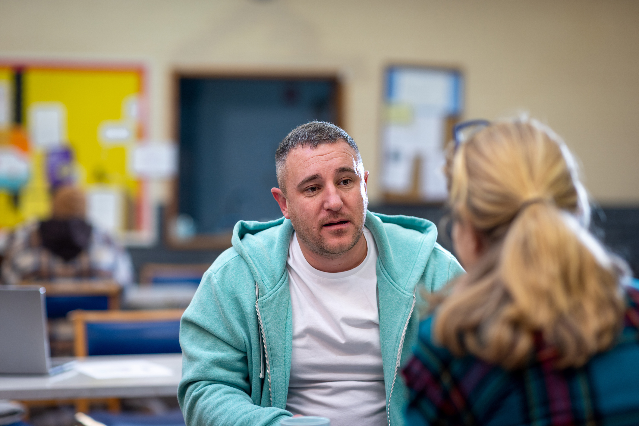 Over-the-shoulder shot of a male adult confiding in a one-to-one with another adult about their mental wellbeing.