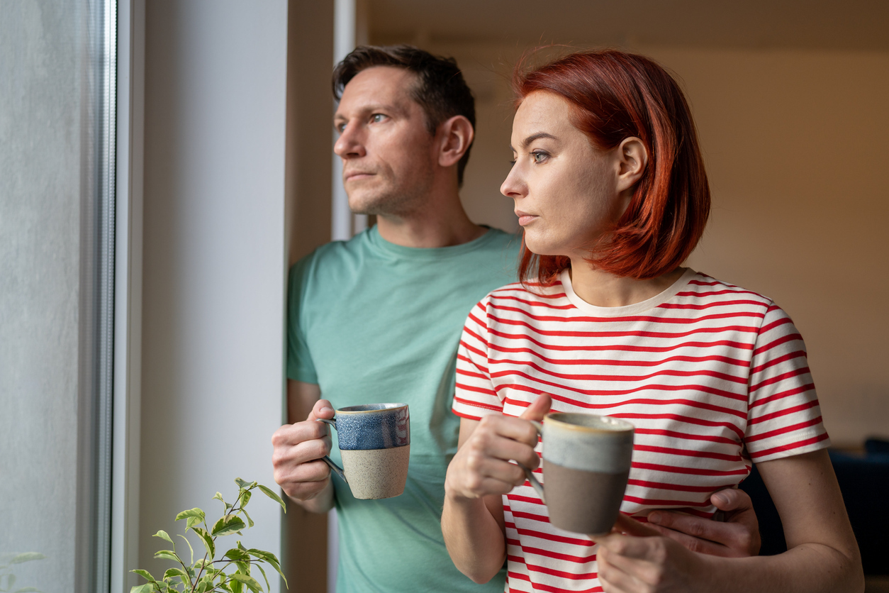 man and a woman stand near each other holding mugs whilst looking pensively out of a window.