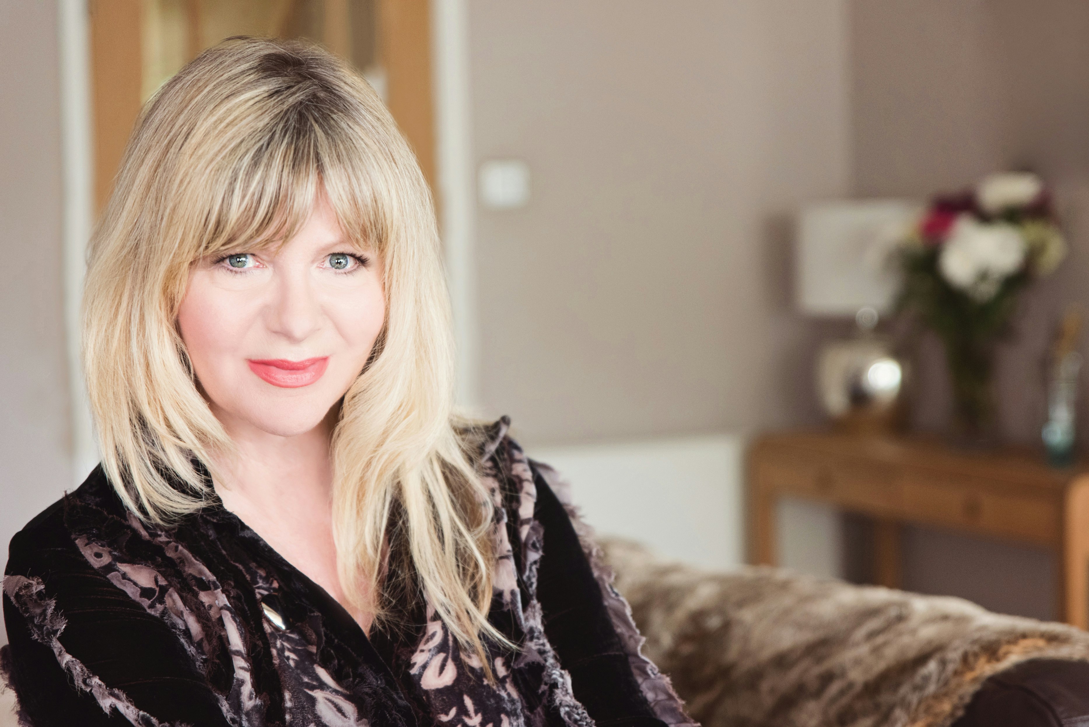 An older woman with blonde smiles from a living room.