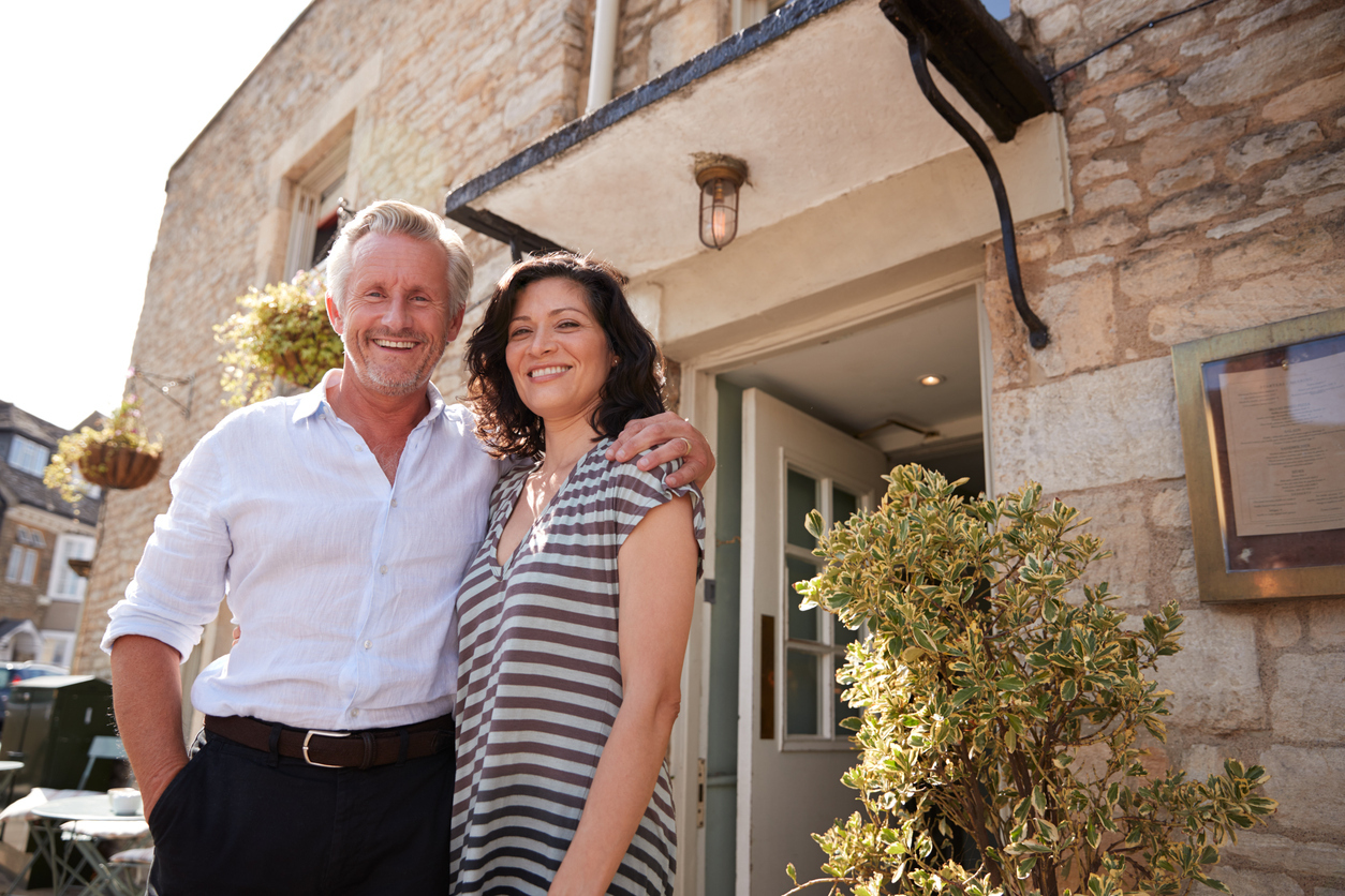 Mature business couple standing outside their restaurant pub smiling in the sunshine