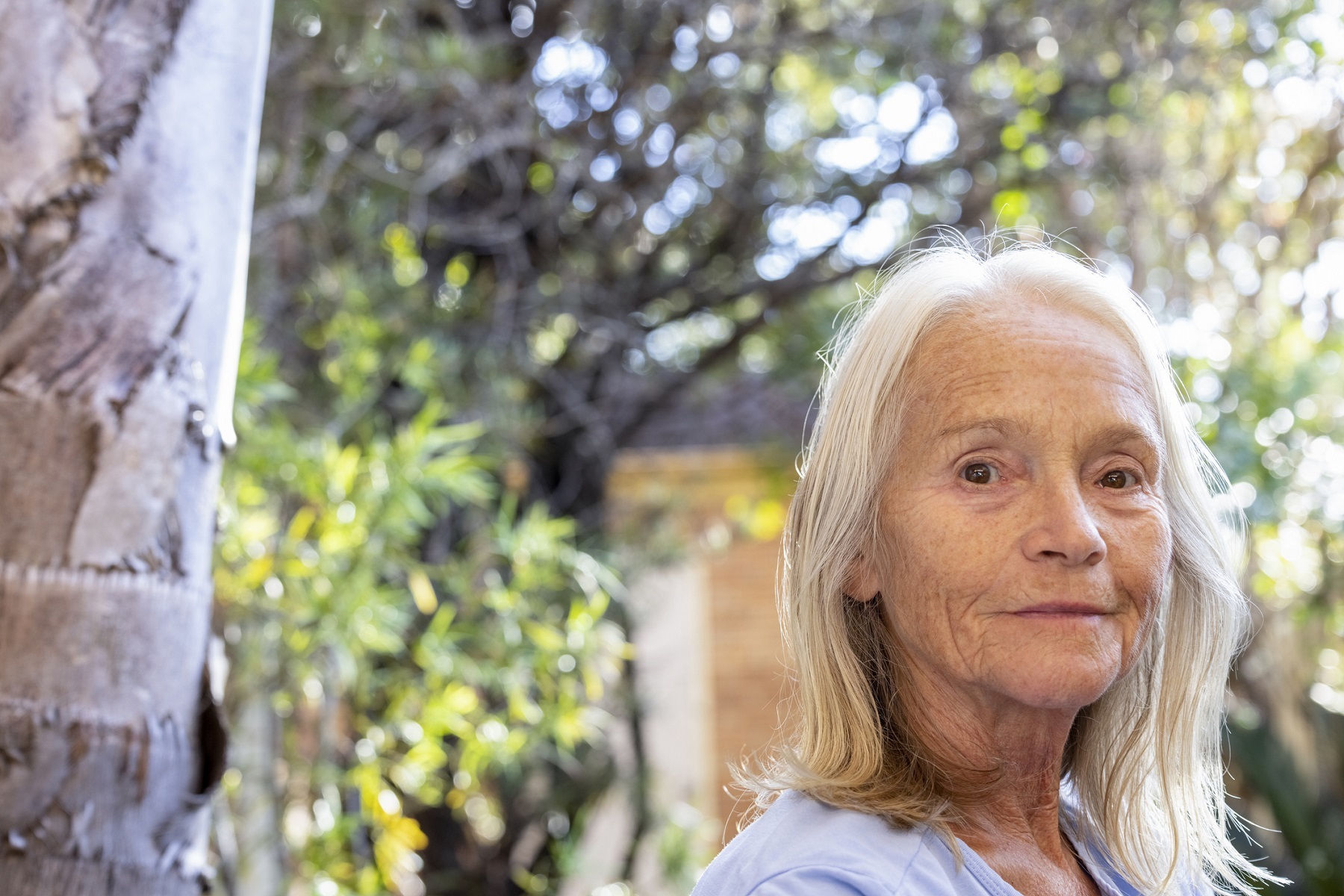 A woman in her mid-sixties with white, shoulder length straight hair turns her head to look at the camera while sitting in a garden.