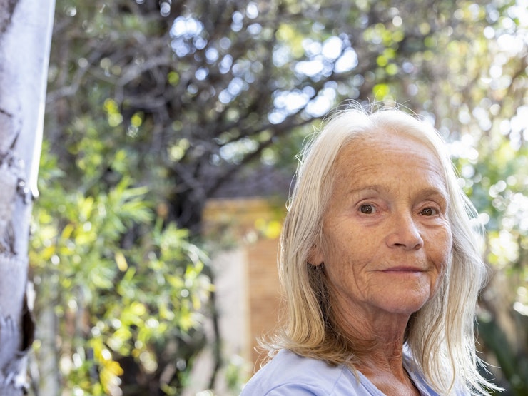 A woman in her mid-sixties with white, shoulder length straight hair turns her head to look at the camera while sitting in a garden.