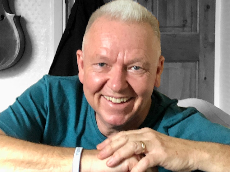 A man in his sixties wearing a blue T-shirt smiles at the camera.