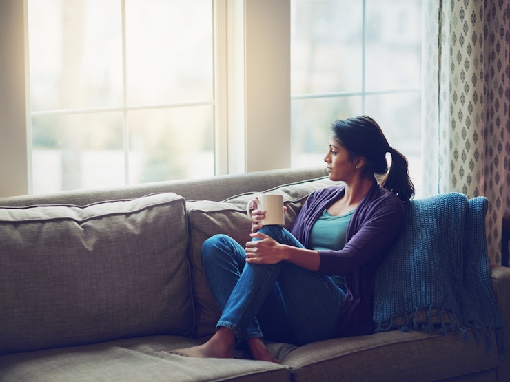 Woman clutching mug on a sofa with her feet up looking thoughtfully out of window.