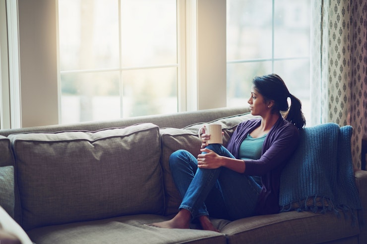 Woman clutching mug on a sofa with her feet up looking thoughtfully out of window.