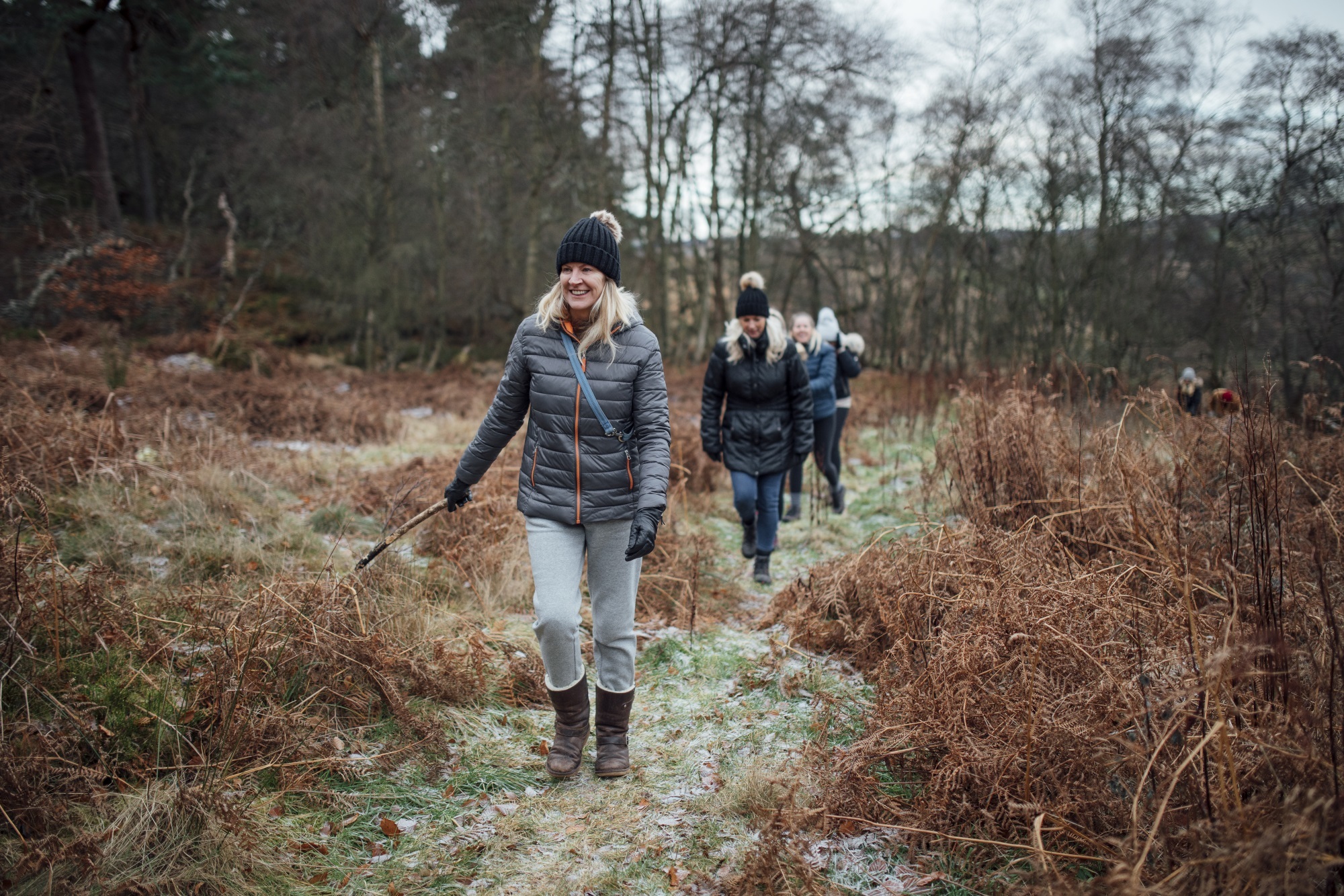 A woman with long blonde hair wearing a wooly hat walks through a wintry forest with others.